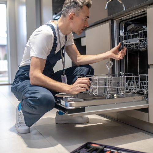 Diagnostics of equipment. Profile of serious man in dark overalls with badge crouching attentively looking at open dishwasher in kitchen at home during day